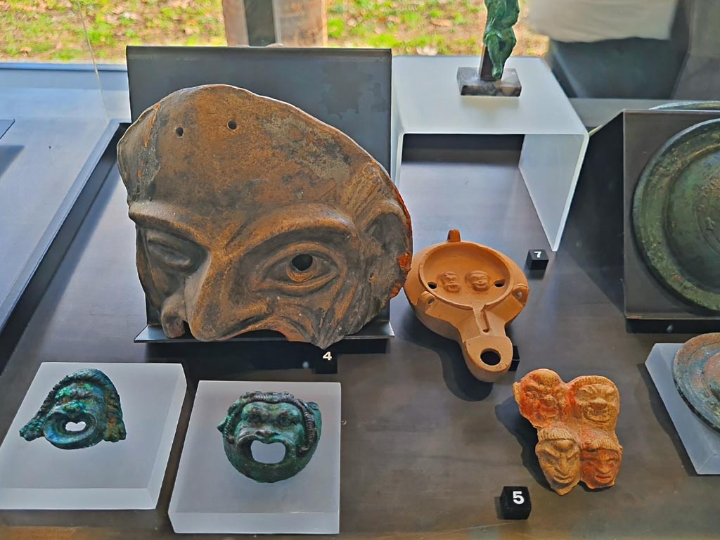 I.9.9 Pompeii. March 2024. No.6 (number hidden, but beneath No.7) – Oil lamp with theatre masks. Photo courtesy of Giuseppe Ciaramella.
On display in exhibition in Palaestra entitled – “L’altra Pompei, vite comuni all’ombra del Vesuvio”.
No.3, lower left – Bronze wall sconces with theatre masks, provenance not known, but from Pompeii. (1st century AD).
No.4 – Terracotta theatre masks Pompeii – provenance not known, but from Pompeii. (1st century AD). 
No.5 – Plaque with theatre masks, (1st century BC – 1st century AD), from I.9.13. 

