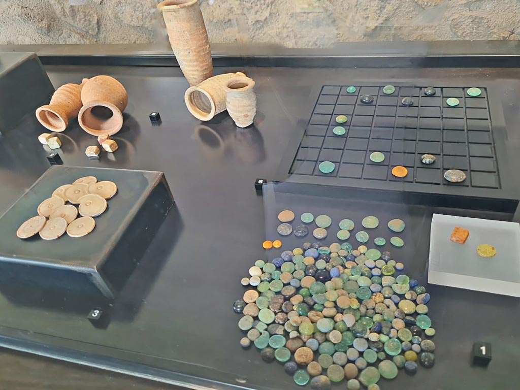 On display in exhibition in Palaestra entitled – “L’altra Pompei, vite comuni all’ombra del Vesuvio”. March 2024.
No.1 – Gaming counters made of glass paste, die and tesserae made of bone, from Porta Stabia Necropolis.
No.2 – Worked bone dice, from V.4.13, Pompeii.
No.3 – Gaming counters made of worked bone, from VI.16.26/27, Pompeii.
No.5 – Pottery dice shakers, from II.1.6, Pompeii.
No.6 – Reproduction of a gaming table. 
Photo courtesy of Giuseppe Ciaramella.
