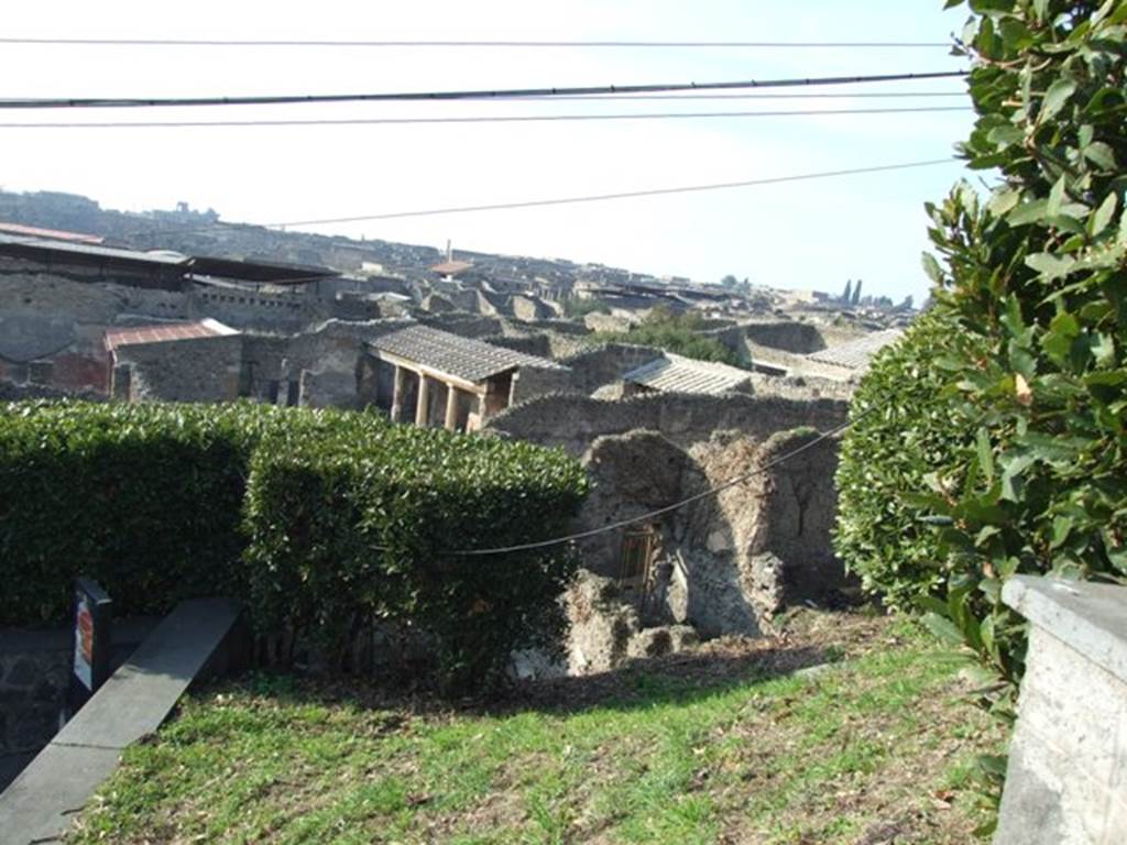 Looking north-west from Casina dell’Aquila to rear of entrance at IX.7.14, lower centre. 1968.
On the left can be seen the west wall of the triclinium of IX.7.12.
The west wall of the garden can be seen behind the greenery, on the right of the doorway.
According to Boyce –
Against the west wall of the garden stands an aedicula, built upon a square masonry base (0.92 square, h.0.70).
The sides of the base are decorated with green plants painted on the white ground.
Above the base, walls enclose a niche (h.0.42, w.0.52, d.0.62).
In front of the structure stands a small altar (0.24 by 0.23, h.0.80).
The roof (no longer preserved) originally extended out over the altar as well as the niche, and the front of it was adorned with a pediment.
It was supported by two columns, the marks of the bases of which are still to be seen near the corners of the base.
On the walls within the niche is the lararium painting.
On the back wall to the right of an altar, stands the Genius pouring a libation upon it.
To the left of the same altar is a serpent, the head of which was done in stucco relief.
On each of the side walls are painted three figures: from back to front – a plant, a Lar, a man in a white garment holding in his left hand an object resembling a black cup, with a patera in his right hand.
He quotes references – Not. Scavi, 1881, 92-93; Bull. Inst., 1883, 146.
In Not. Scavi, it is described as House, only partly excavated – “and corresponds to, after the sixth doorway, west side, counting from the north-west”.
In Bull. Inst, it is described as “West side, first entrance counting from the south, garden in part excavated.”
See Boyce G. K., 1937. Corpus of the Lararia of Pompeii. Rome: MAAR 14. (p.89, no.445).
