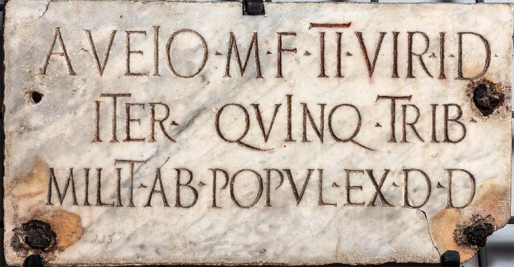 HGW02 Pompeii. On the 16th March 1763 a marble plaque, (20BC – 10AD), was found set in the tufa block on the back of the schola. 
According to Mau the marble plaque had the inscription -

A VEIO M F IIVIR I D
ITER QVINQ TRIB
MILIT AB POPUL EX D D

Now in Naples Archaeological Museum. Inventory number 3887 (CIL X, 996)

According to the information card in the Museum, it read –
“To Aulus Veius, son of Marcus, duovir with judicial powers, the second time with censorial powers, military tribune nominated by the people, by decree of the municipal council”.
(Aulus Veius was a member of the ruling class of the Augustan age.
The office of tribunus militum a populo attests to his achievement of Roman knighthood. 
His family, who had come to Pompeii as Sullan colonists, is no longer attested after him.) 

According to Epigraphik-Datenbank Clauss/Slaby (See www.manfredclauss.de) this read
 
A(ulo) Veio M(arci) f(ilio) IIvir(o) i(ure) d(icundo)
iter(um) quinq(uennali) trib(uno)
milit(um) ab(!) popul(o) ex d(ecreto) d(ecurionum)      [CIL X 996]

According to Cooley this translates as 

To Aulus Veius, son of Marcus, duumvir with judicial power twice, quinquennial, military tribune by popular demand. By decree of the town councillors.

See Kockel V., 1983. Die Grabbauten vor dem Herkulaner Tor in Pompeji. Mainz: von Zabern. (p. 51).
See Cooley, A. and M.G.L., 2004. Pompeii: A Sourcebook. London: Routledge. (p. 139, G4).



